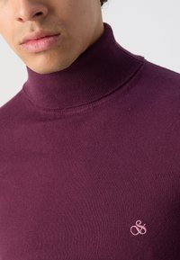 Pull à col roulé bordeaux en tissu doux, avec un col côtelé et un petit logo rose sur la poitrine. Texture lisse.