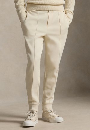 Krēmkrāsas piegriezuma sporta bikses ar sānu krokām, ribotām aprocēm un priekšējiem kabatām, kombinētas ar gaiši bēšām sporta kurpēm. Gluda tekstūra.