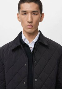 Chaqueta acolchada negra con cuello, que presenta botones a presión plateados y una textura suave, llevada sobre un suéter oscuro y una camisa blanca.