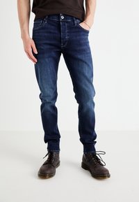 Jeans in denim blu scuro slim-fit con finitura sbiadita, cinque tasche e zip, abbinati a stivaletti grigi con lacci alla caviglia.