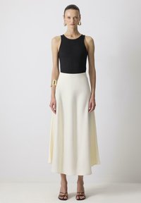 Zwarte ribgebreide mouwloze top gecombineerd met een vloeiende crèmekleurige maxi-rok. Accessoires omvatten een gouden armband om de linkerpols en zwarte sandaaltjes met banden.