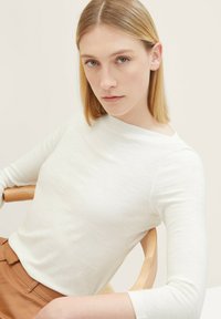 Un haut à manches longues blanc avec un col montant et une texture douce, associé à un pantalon marron taille haute, présenté sur un fond uni.