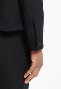 Svart skräddarsydd jackärm med knappmanschett, visas tillsammans med en hand. Tygets struktur ser slät ut, med en strukturerad design.