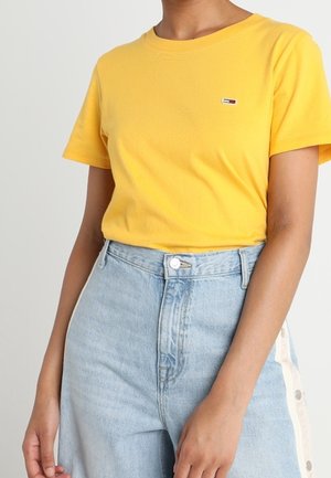 Henkilö, jolla on keltainen lyhythihainen paita, jossa pieni logo, ja vaaleansiniset korkeavyötäröiset farkut, joissa on nappi- ja vetoketjusulku.