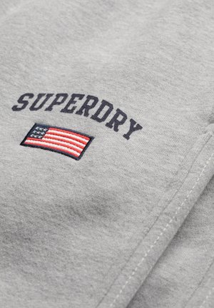 Sudadera gris con "SUPERDRY" en texto negro sobre un diseño bordado de la bandera estadounidense. La textura parece suave y lisa.