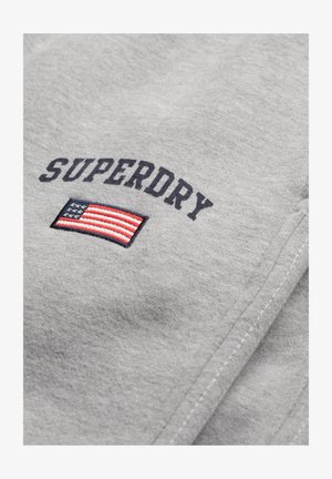 Sudadera gris con "SUPERDRY" en texto negro sobre un diseño bordado de la bandera estadounidense. La textura parece suave y lisa.