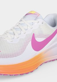 Λευκό αθλητικό παπούτσι Nike με ροζ σήμα, λευκά κορδόνια και πορτοκαλί σόλα με διαβάθμιση σε ανοιχτό φόντο.
