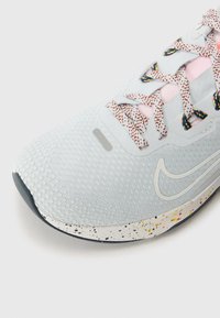 Chaussure de sport gris clair avec tige en mesh texturé, languette rose, lacets à motifs et semelle blanche tachetée avec un swoosh Nike blanc.