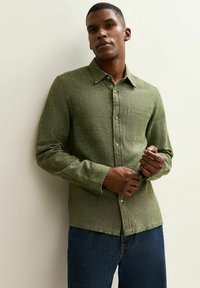 Chemise boutonnée verte texturée avec col, poche poitrine simple et boutons en bois, portée sur un pantalon en denim bleu.