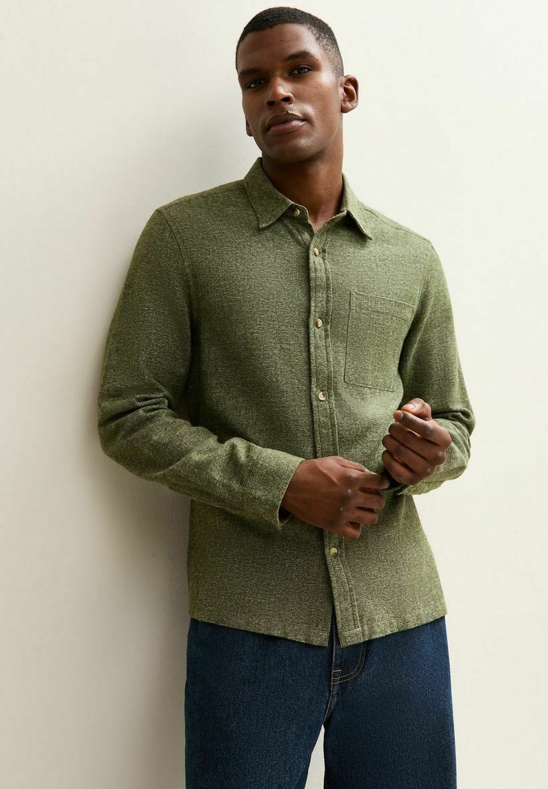 Chemise boutonnée verte texturée avec col, poche poitrine simple et boutons en bois, portée sur un pantalon en denim bleu.