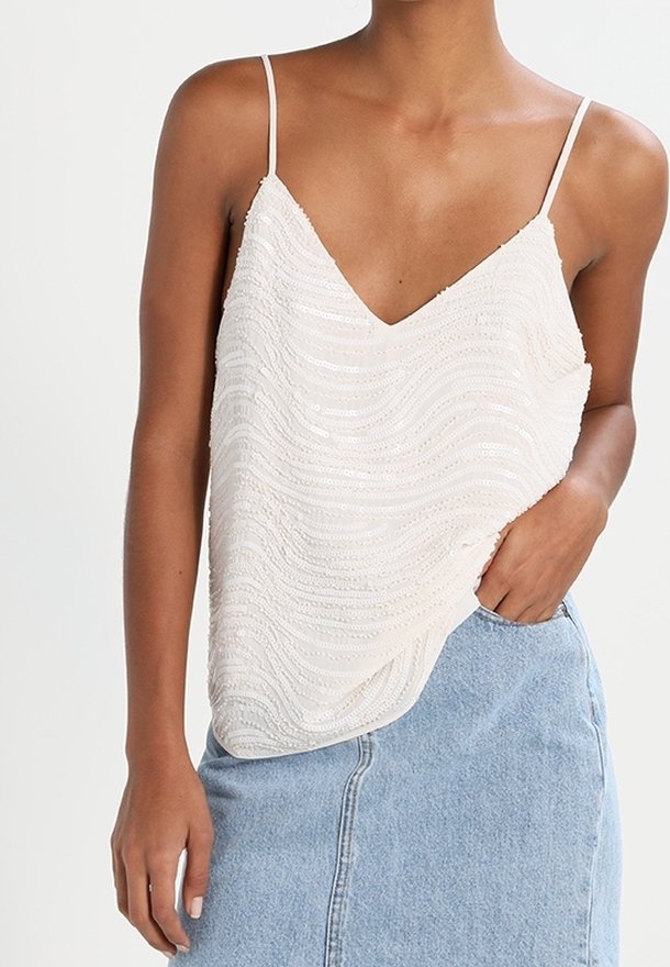 Femme portant un haut sans manches blanc à sequins avec de fines bretelles et une jupe en denim bleu clair, une main dans la poche.
