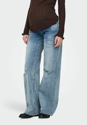 Gravid person iført slitte vide blå jeans, en tettsittende mørkebrun langermet topp og svarte sko, stående mot en ensfarget bakgrunn.