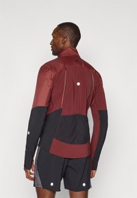 Giacca atletica bordeaux e nera con maniche lunghe, dotata di accenti riflettenti, tasche con zip e colletto alto. Materiale leggero.
