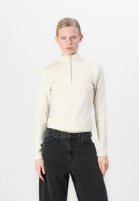 Kremasti rebrasti long-sleeve top s visokim ovratom i polu-zamkom, u kombinaciji s crnim trapericama visokog struka. Glatka tekstura i uski kroj.