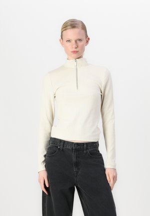Cream ribbed long-sleeve top with a high collar and half-zip, paired with high-waisted black denim jeans. Smooth texture and fitted design.