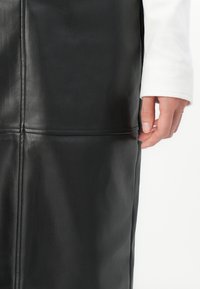 Gros plan d’une main à côté des coutures verticale et horizontale sur une jupe en cuir noire, avec une manche blanche partiellement visible.