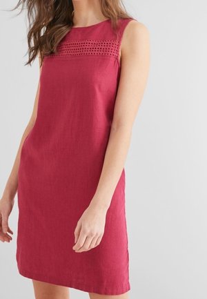 Kvinde iført ærmeløs, knælængde rød kjole med blonder over brystet, stående foran en ensartet lys grå baggrund.