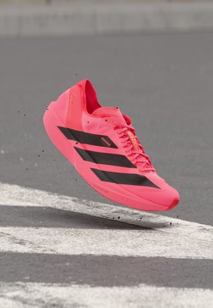 Zapatilla de correr de color rosa brillante con rayas negras, cordones y texto "Adizero", capturada en el aire sobre una superficie de asfalto gris con marcas blancas.