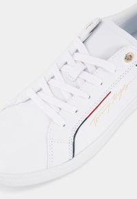 Baskets en cuir blanc avec lacets blancs, œillet doré et logo Tommy Hilfiger en écriture dorée avec des accents de design rouges et bleu marine.