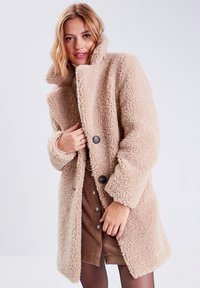 Cappotto teddy beige con colletto alto, realizzato in tessuto testurizzato. Presenta bottoni scuri e una vestibilità comoda, con dettagli di cucitura visibili.