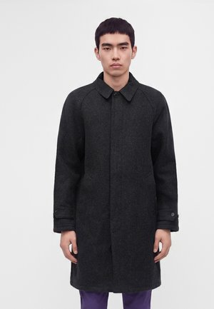 Jeune homme portant un long manteau gris foncé et un pantalon violet, debout devant un fond uni gris clair.