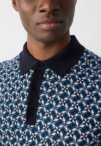 Sweter polo z dzianiny w geometryczny wzór w kolorach niebieskim, czarnym i białym. Posiada jednolitą granatową kołnierz i zapięcie na trzy guziki.