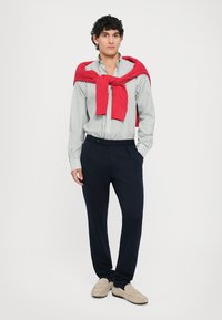 Dryžuotas balto ir mėlyno švarkelis, raudona megztinė surišta aplink pečius, tamsiai mėlynos kelnės, šviesūs slip-on batai, neutralus fonas.