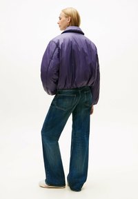 Giacca a vento viola con colletto alto, abbinata a jeans in denim scuro a vestibilità ampia. La texture della giacca è liscia e lucida.