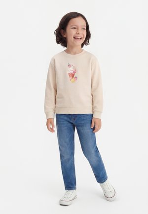 Usmievavé dieťa s tmavými vlasmi, ktoré má na sebe béžovú mikinu s motívom zmrzlinového kornúta, modré džínsy a biele tenisky, stojí na bielom pozadí.