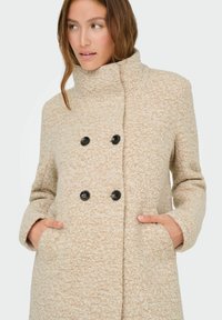 Manteau beige croisé en mélange de laine texturée, avec des boutons noirs, un col haut et des poches latérales.