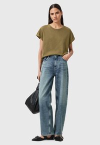 Olivgrünes Baumwoll-T-Shirt mit aufgerollten Ärmeln, kombiniert mit hellblauen weiten Jeans. Schwarze flache Schuhe und eine schwarze Tasche runden das Outfit ab.