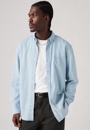 Chemise en denim bleu clair, associée à un t-shirt blanc à col rond et une ceinture noire, portée sur un pantalon sombre.