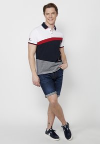 Cămășuța polo pentru bărbați în alb, bleumarin și gri cu accente roșii; mâneci scurte, design cu guler, asociată cu pantaloni scurți din denim rulați și adidași bleumarin.
