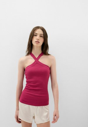 Vrouw met lang bruin haar draagt een mouwloze frambozenkleurige haltertop en lichtbeige short, staand tegen een effen witte achtergrond.