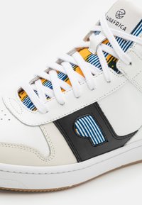 Fehér bőr sportcipő, fekete díszítéssel és mintás textilel a nyelvén, kék és sárga csíkokkal. Fehér fűzők és gumitalp.