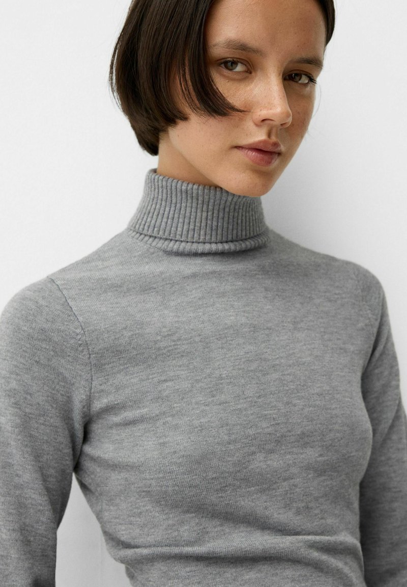 Junge Frau mit kurzem braunem Haar, die einen grauen, gerippten Rollkragenpullover trägt und leicht zur Seite schaut, vor einem schlichten weißen Hintergrund.