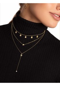 Collier en or avec plusieurs chaînes, pendentifs circulaires, une lettre "M" et un accent en forme d'étoile. Porté sur un col roulé noir.