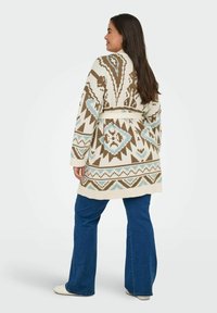 Ženska z dolgimi temnimi lasmi, oblečena v vzorčasto bež, rjavo in modro pleteno jopico, modre razširjene kavbojke in bele čevlje, obrača se stran.