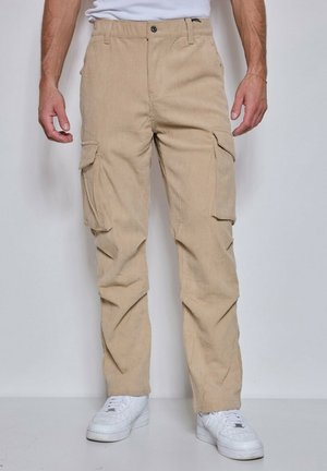 Pantalon cargo en velours côtelé beige avec poches à rabat porté par une personne debout, associé à des baskets blanches et une chemise blanche rentrée.