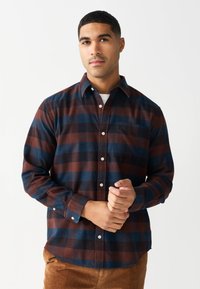 Koszula w kratę z długim rękawem w kolorach ciemnobrązowym, niebieskim i czerwonym. Wykonana z flaneli, posiada zapięcie na guziki z przodu, kieszeń na piersi oraz regulowane mankiety.