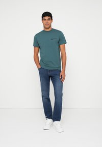Teal cotton t-shirt with a crew neck and small logo. Paired with blue denim jeans and white sneakers. Simple design and relaxed fit.