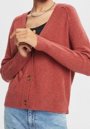 Nainen, joka käyttää ruosteenväristä napitettavaa neuletakkia mustan topin ja vaaleansinisten farkkujen päällä, hopeaketjukaulakorun kanssa.