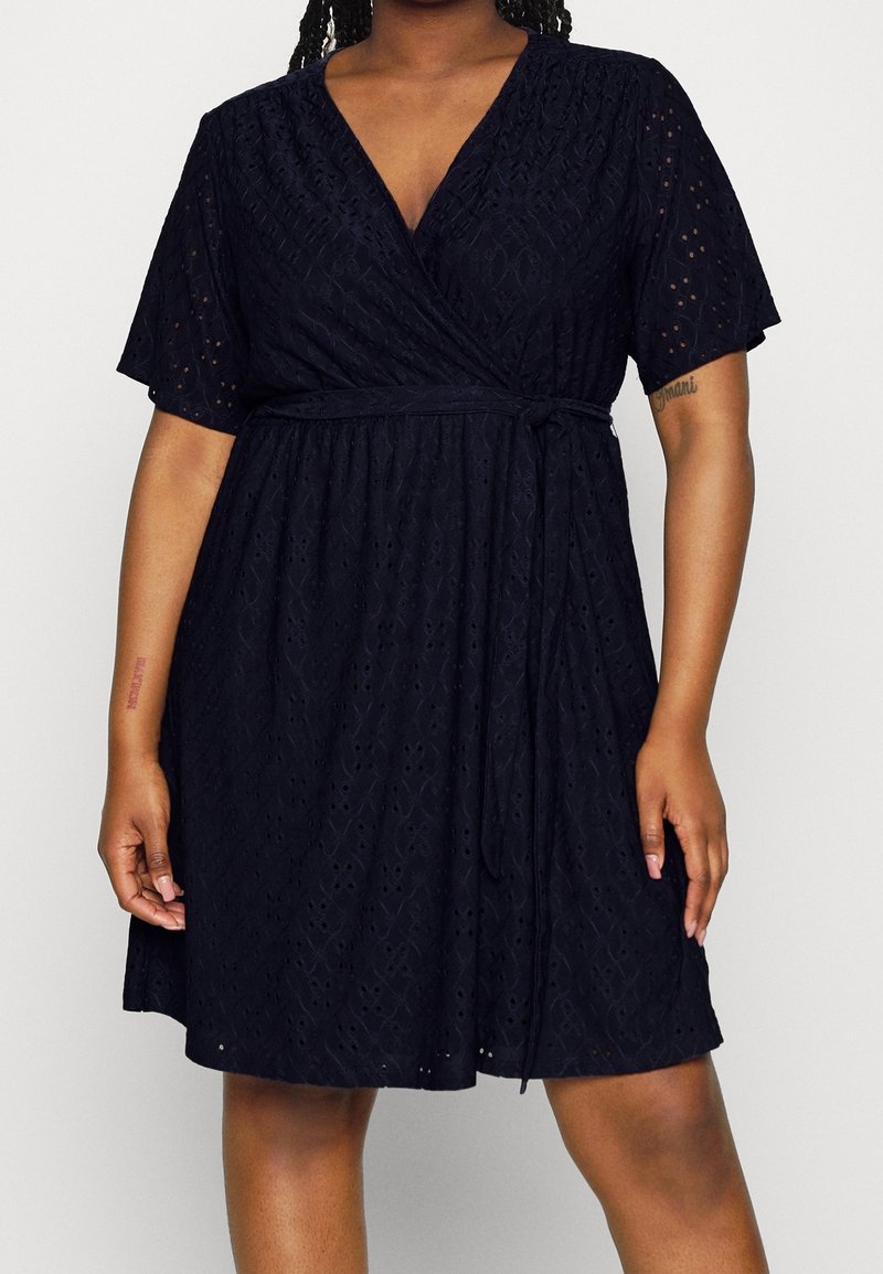 Nainen, joka on pukeutunut polvipituiseen tummansiniseen kietaisumekkoon, jossa on lyhyet hihat ja solmittava vyö, paljastaen osittaisia tatuointeja käsissään.