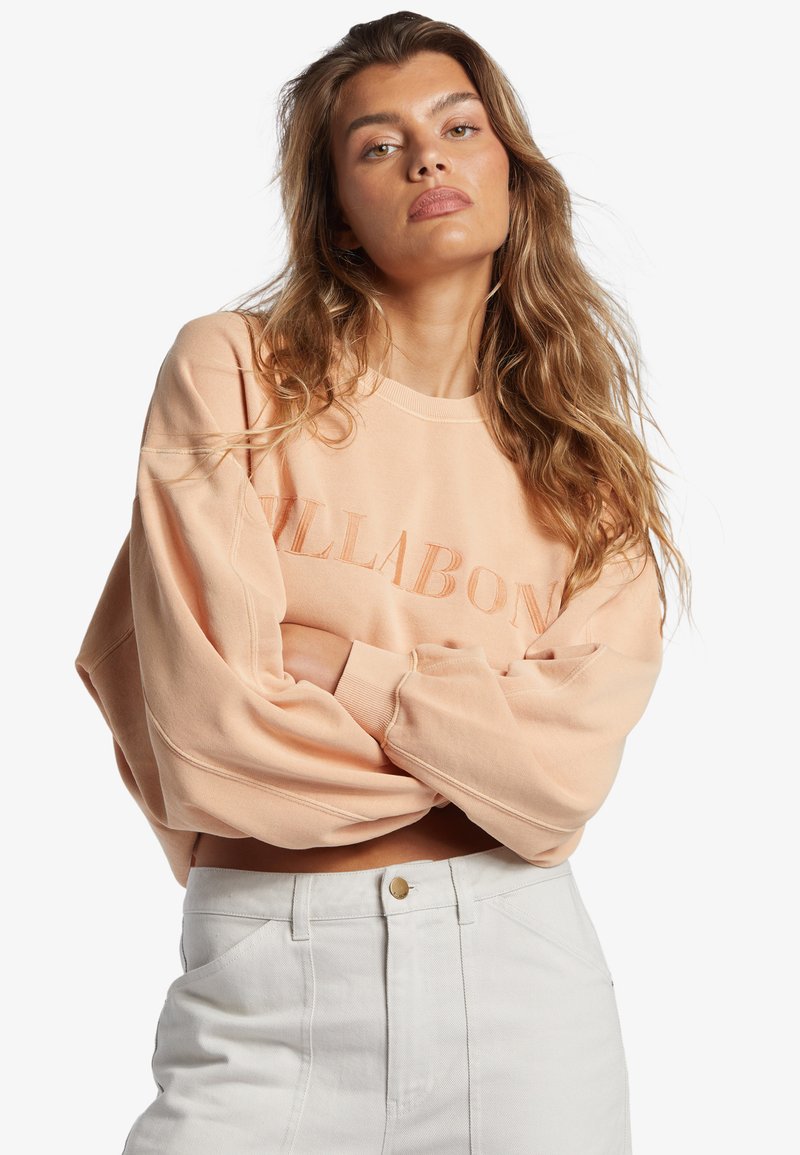 Svetloružová oversized mikina s vyšitým nápisom "BILLABONG", s padnutými ramenami a rebrovanými manžetami, kombinovaná s bielymi rifľami so zvýšeným pásom.