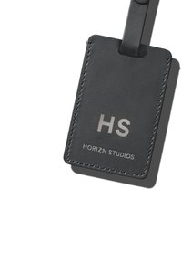 Étiquette de bagage en cuir noir avec des initiales embossées "HS" et "HORIZN STUDIOS", finie avec une couture contrastante et un attache en boucle.