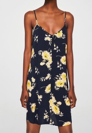 Robe bleu marine sans manches avec fines bretelles et motif floral jaune portée par une personne debout sur un fond uni.