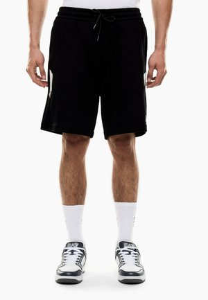 Mann trägt schwarze Shorts mit Kordelzug, weiße Sportsocken und weiße sowie schwarze Schnürsneaker, steht vor weißem Hintergrund.