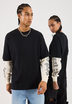 To personer med fletninger bærer lagdelte sort og creme grafiske langærmede skjorter, den ene sat sammen med lys blå jeans, den anden med slidte sorte bukser.
