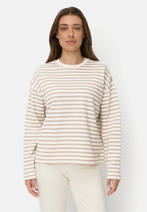 Kvinde iført en beige og hvid stribet langærmet skjorte og lyse bukser står mod en ensfarvet hvid baggrund.