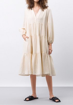 Woman standing in a loose, tiered cream midi dress with long sleeves and black flat slide sandals on a gray floor.